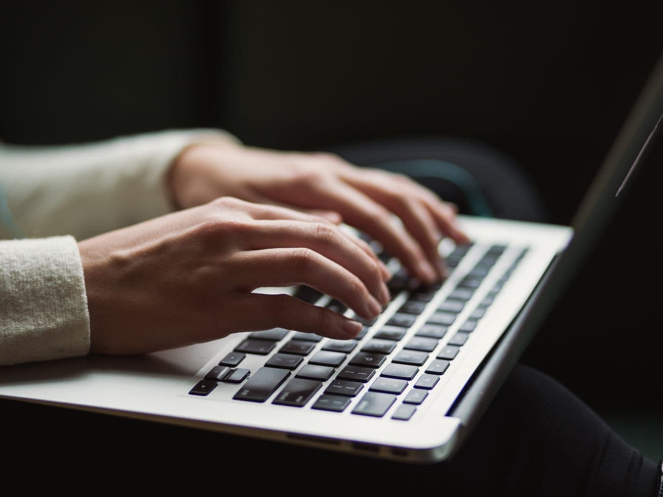 Close-up of hands in beige sweater sleeve typing on a silver laptop keyboard against dark background, showing modern workplace technology usage