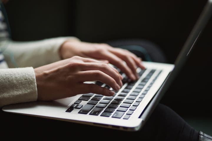 Close-up of hands in beige sweater sleeve typing on a silver laptop keyboard against dark background, showing modern workplace technology usage