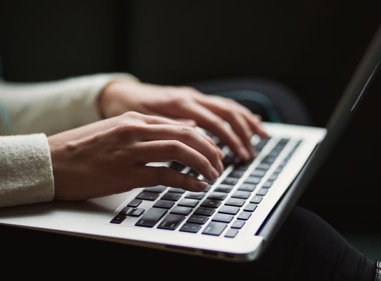 Close-up of hands in beige sweater sleeve typing on a silver laptop keyboard against dark background, showing modern workplace technology usage