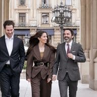 Three people are walking through an archway, talking to each other; in the background, an old apartment building can be seen.