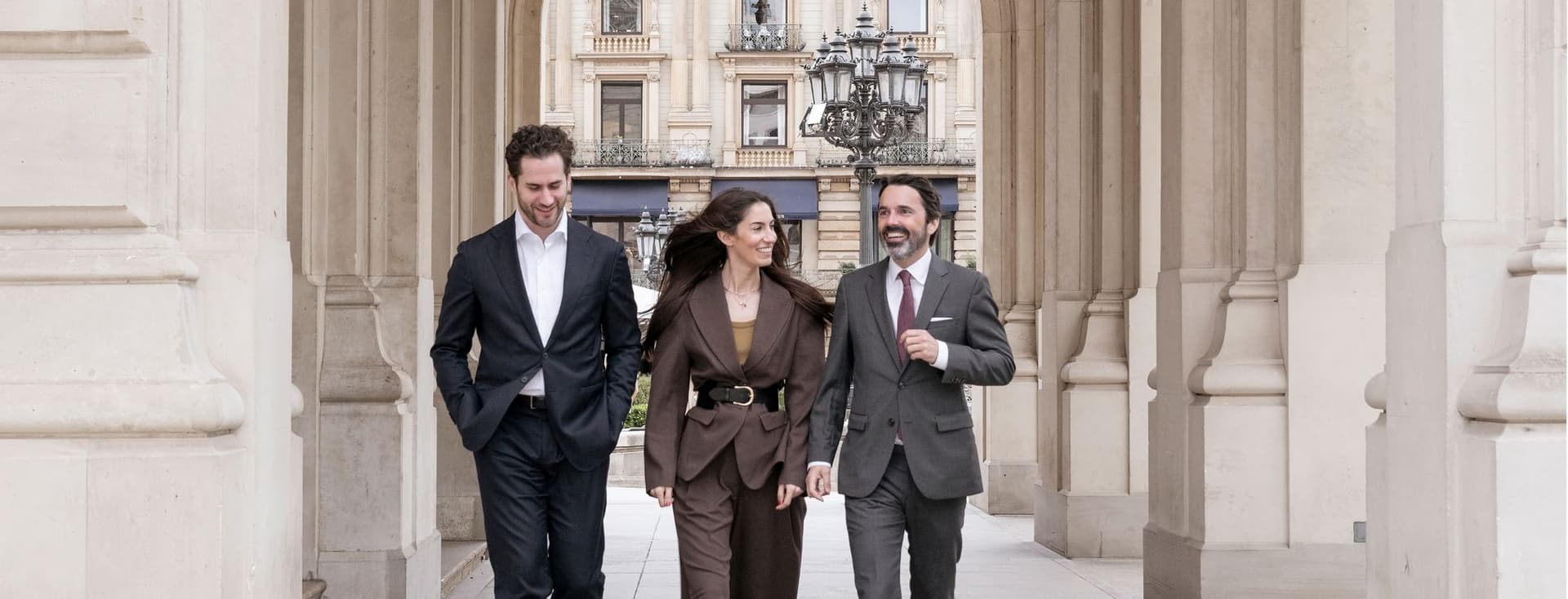 Three people are walking through an archway, talking to each other; in the background, an old apartment building can be seen.