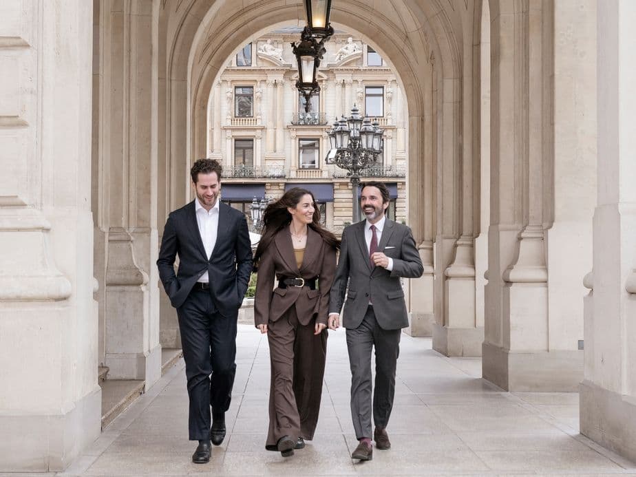 Three people walk through an arcade while chatting, with an old apartment building visible in the background.