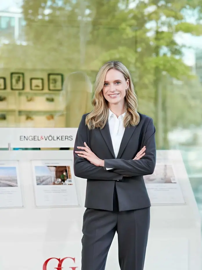 A woman in a black suit and white blouse stands smiling in front of a shop window