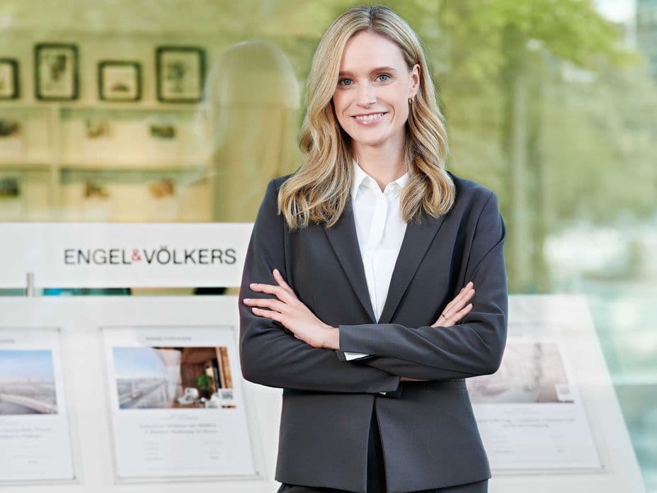 A woman in a black suit and white blouse stands smiling in front of a shop window