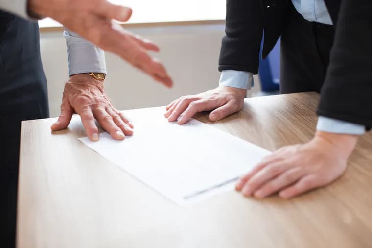 Two people leaning over a wooden table with a document: one has a hand resting on the paper, while the other reaches towards it.