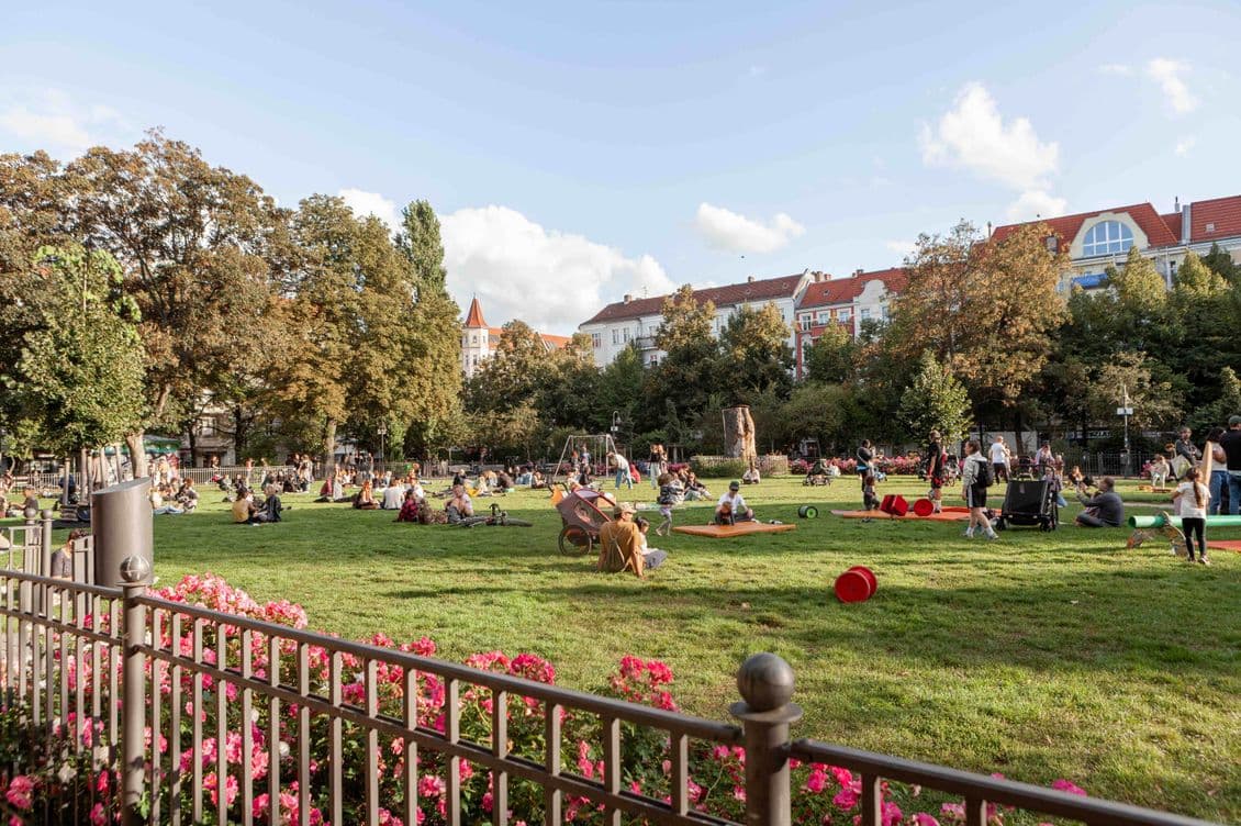People relaxing and enjoying a sunny day in a park with green grass, trees, and a playground. Pink flowers line a fence in the foreground.