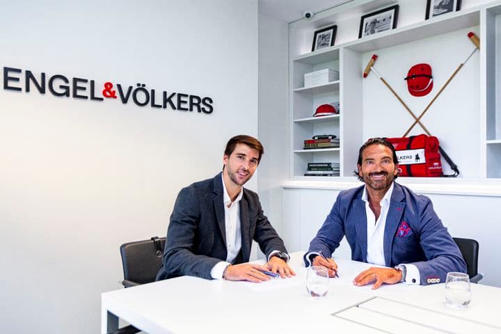 Dos agentes inmobiliarios sonrientes sentados a una mesa blanca en una oficina de Engel & Völkers, firmando documentos. Equipamiento de polo decora las estanterías.