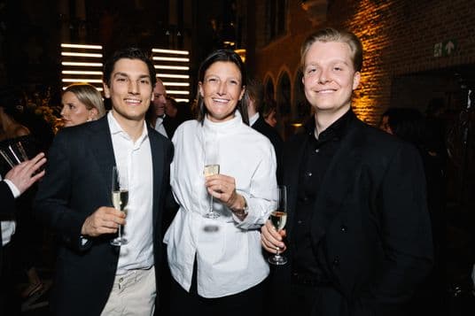 Three well-dressed guests smile for the camera, each holding a champagne flute at a warmly lit brick-walled party.