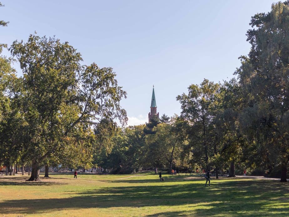 A park with green grass and tall trees, featuring a distant church steeple under a clear blue sky.