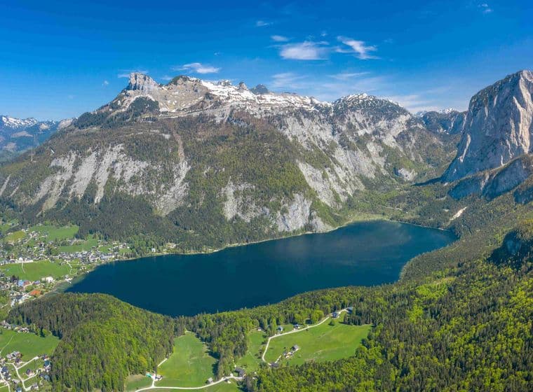 View of Hallstatt, a picturesque village in Austria's Ausseerland region, featuring traditional wooden Alpine houses cascading down the mountainside to Lake Hallstatt. The iconic church spire and historic waterfront buildings are perfectly mirrored in the tranquil lake waters, with majestic mountains forming a dramatic backdrop. This UNESCO World Heritage site represents the quintessential Austrian lakeside property market.