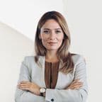 Una mujer rubia con una chaqueta gris está de pie con los brazos cruzados, sonriendo levemente.