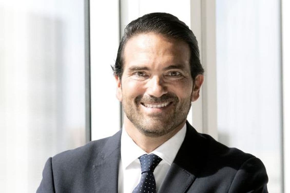 A smiling man in a navy suit and tie stands in front of a window.