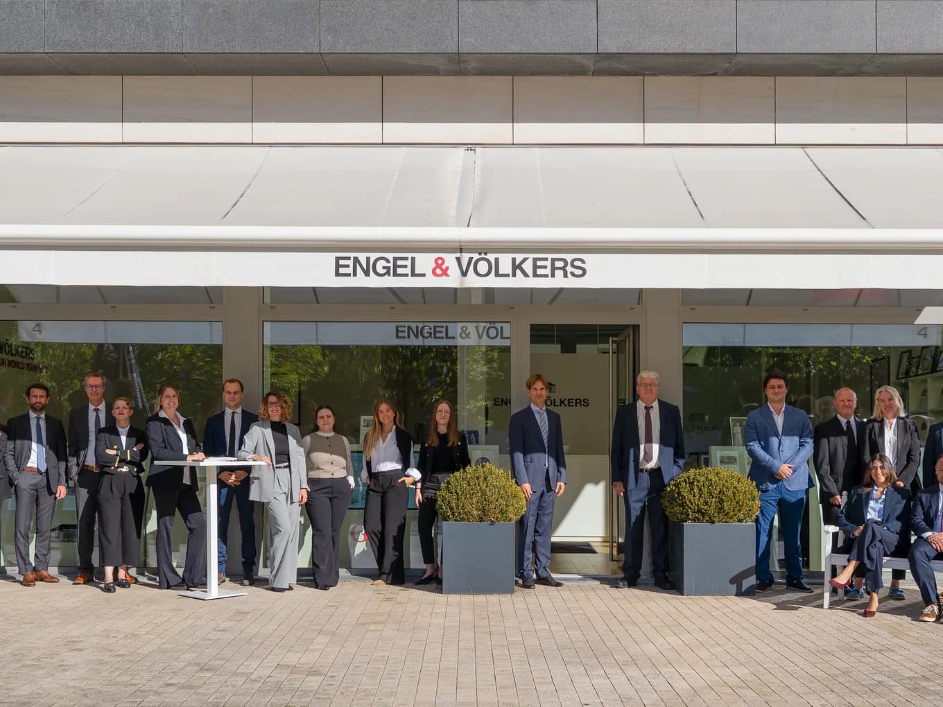 Group of professionals in business attire posing outside an Engel & Völkers real estate office under a white canopy.