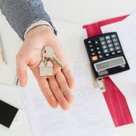 Sheets with numbers and tax calculations next to a calculator and house keys, representing the costs and taxes associated with the purchase of a second home.