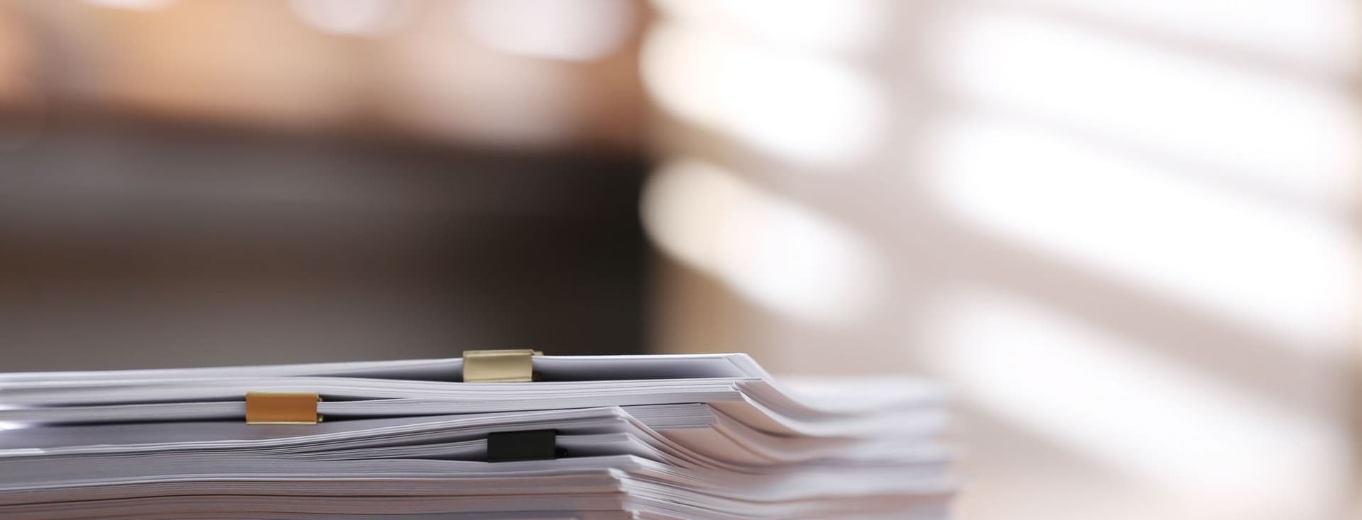 A stack of documents with paper clips on a wooden desk, illuminated by soft, diffused sunlight through blinds in the background.