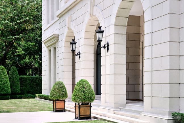 Exterior view of a luxurious white stone building with classical columns, arched entryway, ornate black lanterns, and manicured topiary plants in wooden planters, bordered by a well-maintained garden