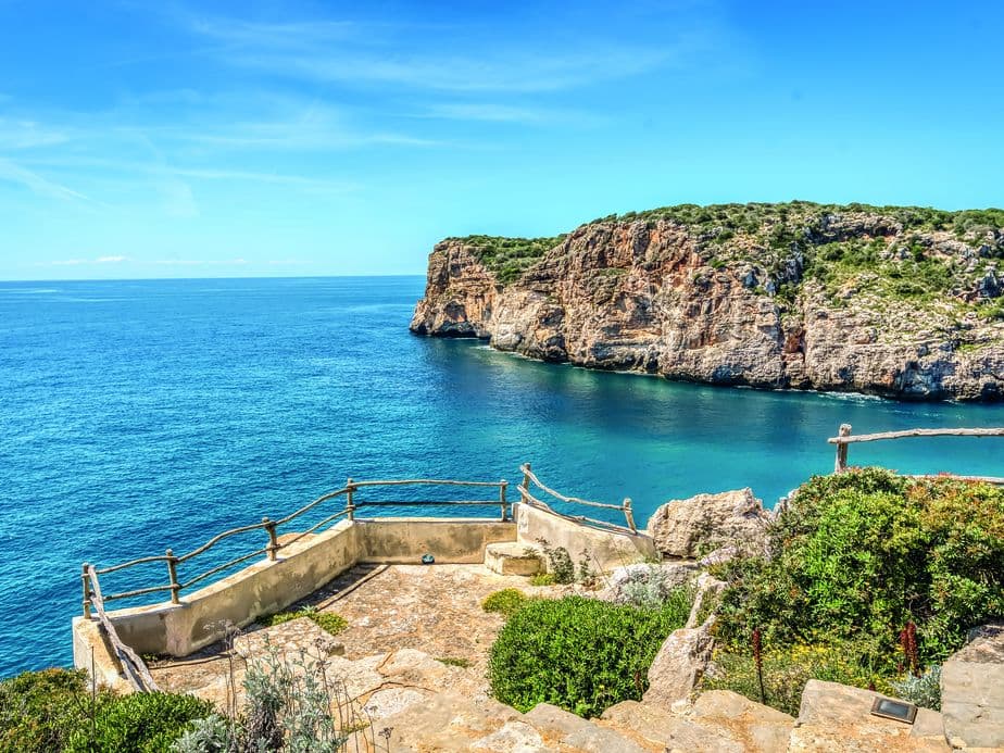 Mirador sobre penya-segats al centre de Menorca amb terrasses de pedra rústiques i baranes de fusta, amb vista al mar turquesa i als impressionants penya-segats de calcària sota un cel blau.