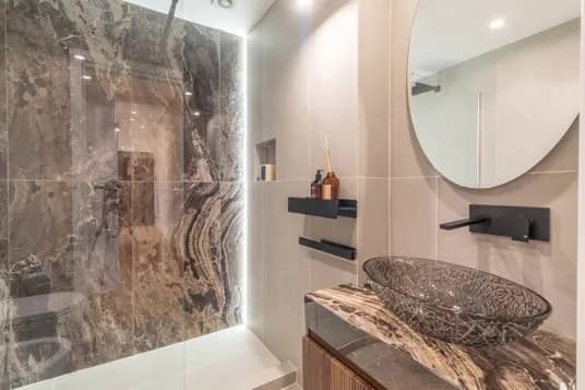 Elegant bathroom with dark marble shower walls, a glass shower enclosure, a round textured vessel sink on a matching marble countertop and a round mirror.