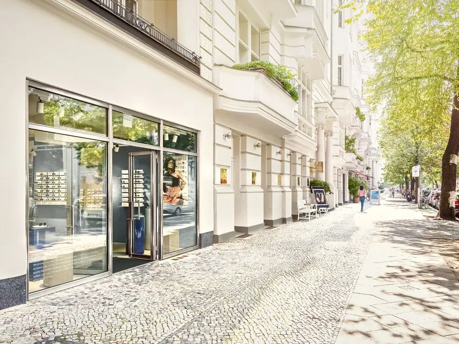 A bright, sunny street view of white buildings with a storefront window displaying eyewear. A woman walks along the sidewalk.