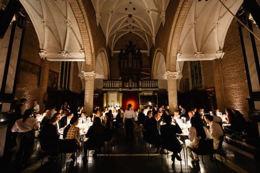 Dimly lit church hall with long candlelit tables, guests in formal attire, Gothic arches and a grand organ at the back.