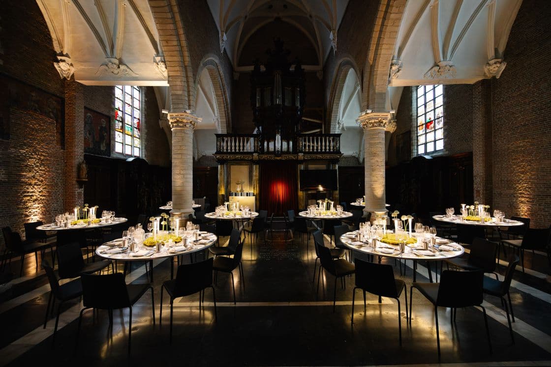Historic church interior set for a banquet: brick walls, white vaulted arches, stained-glass windows, a central organ, and round tables with candles and glassware.