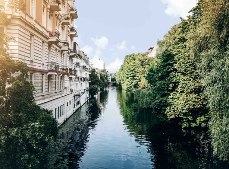 A tranquil canal, lined with lush green trees in Eppendorf and classic European buildings under a clear blue sky.