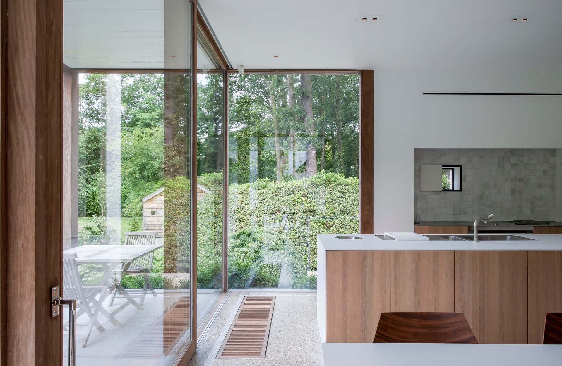 A modern kitchen with wood cabinets and a white countertop, a large window overlooks a green garden with a white table and chairs.