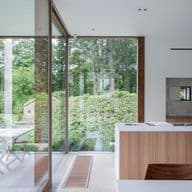 Cucina moderna con accenti in legno, grandi porte in vetro e vista su un giardino lussureggiante. Patio esterno con tavolo e sedie visibile attraverso il vetro.
