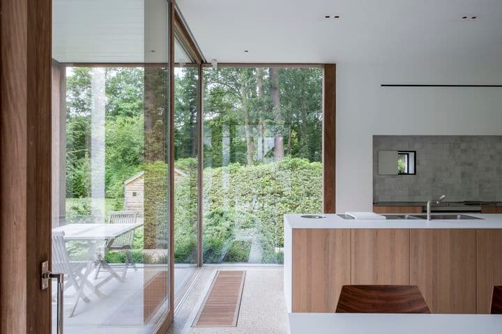 Cucina moderna con accenti in legno, grandi porte in vetro e vista su un giardino lussureggiante. Patio esterno con tavolo e sedie visibile attraverso il vetro.