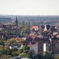 Aerial view of a Hannover with red-roofed buildings, a prominent church tower, and surrounding trees under a clear sky.