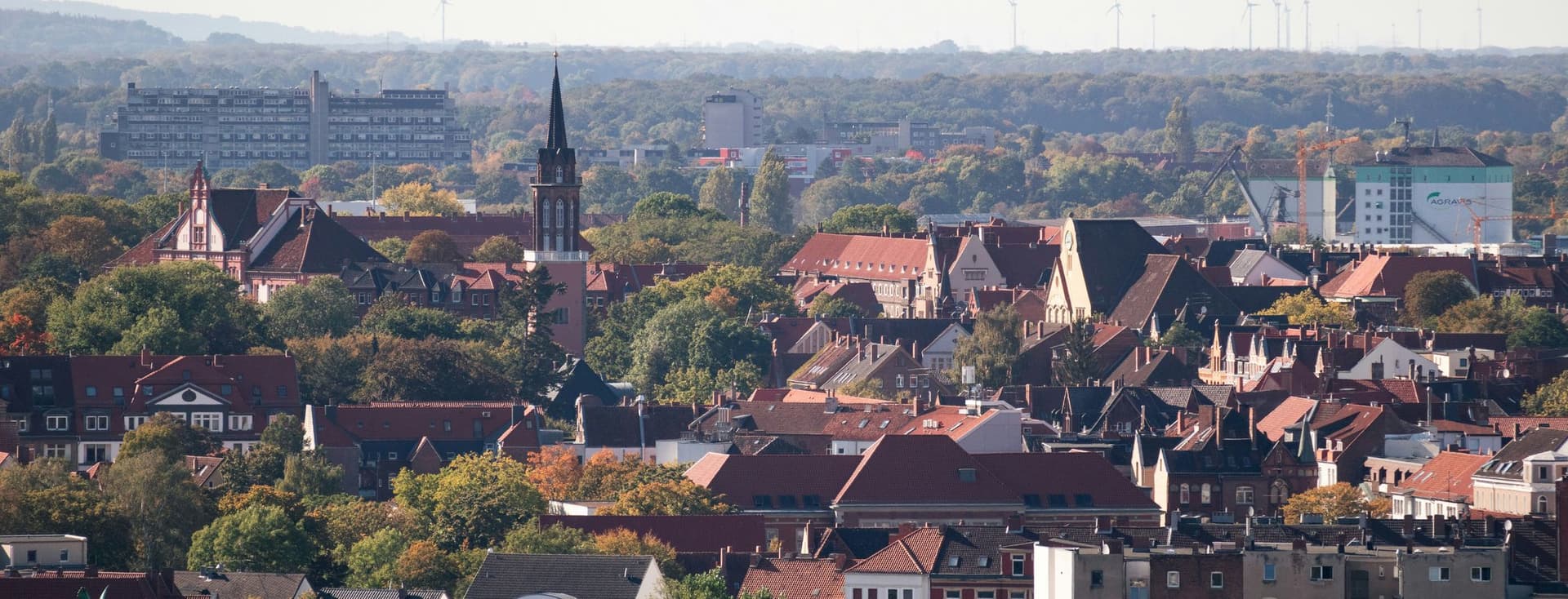 Aerial view of a Hannover with red-roofed buildings, a prominent church tower, and surrounding trees under a clear sky.