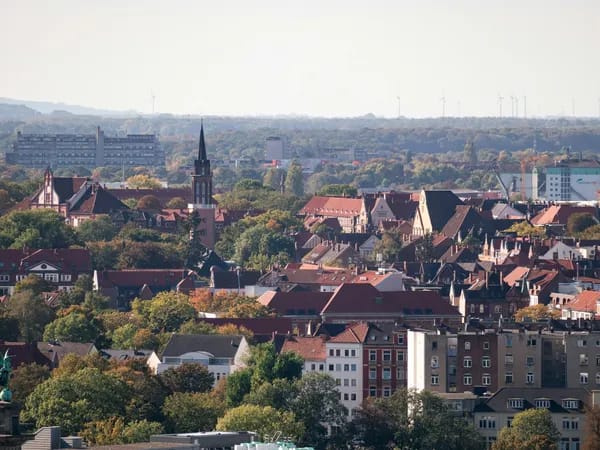 Aerial view of a Hannover with red-roofed buildings, a prominent church tower, and surrounding trees under a clear sky.