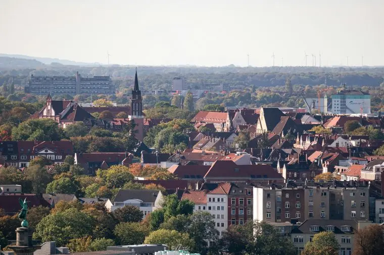 Aerial view of a Hannover with red-roofed buildings, a prominent church tower, and surrounding trees under a clear sky.