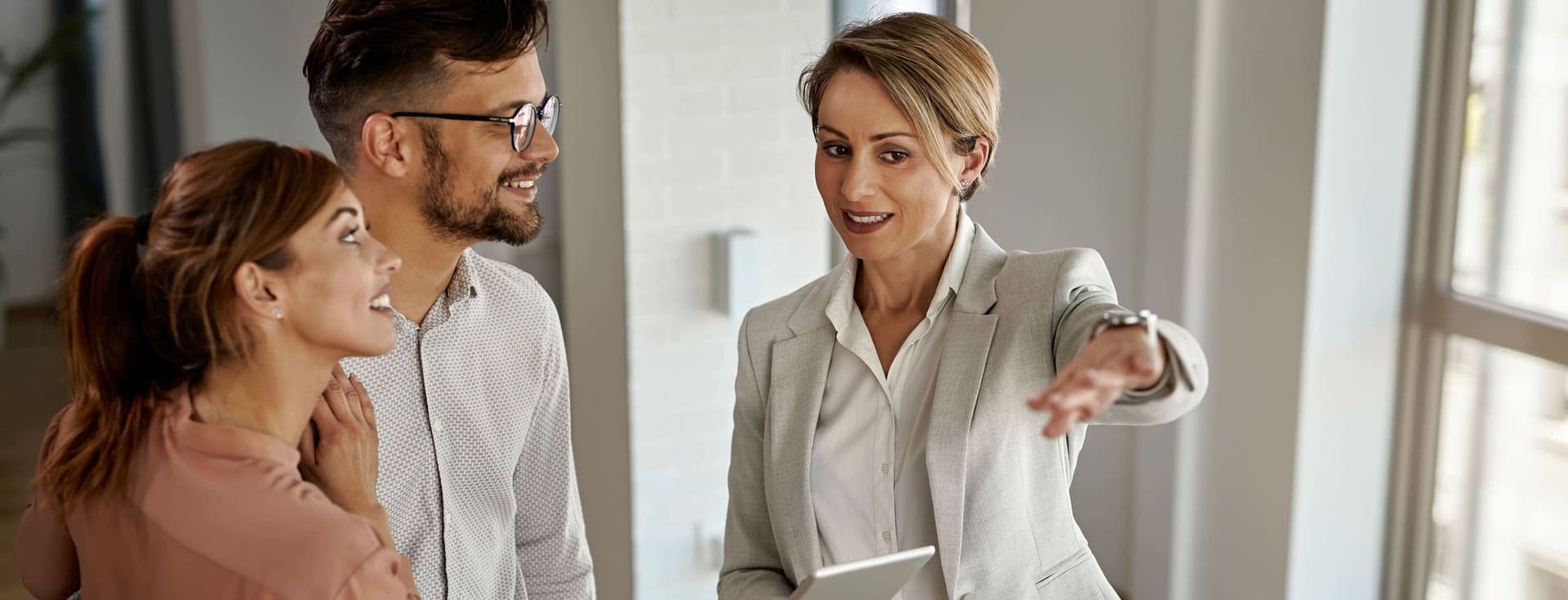 Un agente immobiliare in un abito grigio indica una coppia in uno spazio luminoso e moderno. Tiene in mano un tablet e sorride.