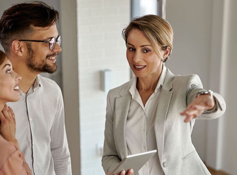 Un agente immobiliare in un abito grigio indica una coppia in uno spazio luminoso e moderno. Tiene in mano un tablet e sorride.