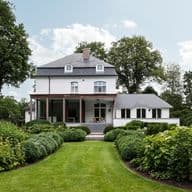 Rear view of a luxurious white estate with a manicured green lawn, meticulously designed boxwood hedges, and professional landscaping under a bright sky.