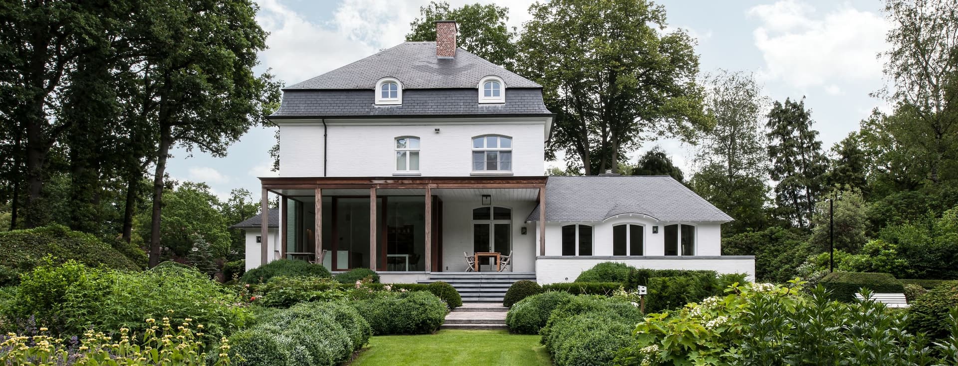 Rear view of a luxurious white estate with a manicured green lawn, meticulously designed boxwood hedges, and professional landscaping under a bright sky.