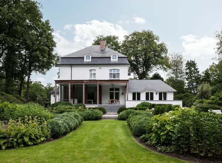 Rear view of a luxurious white estate with a manicured green lawn, meticulously designed boxwood hedges, and professional landscaping under a bright sky.