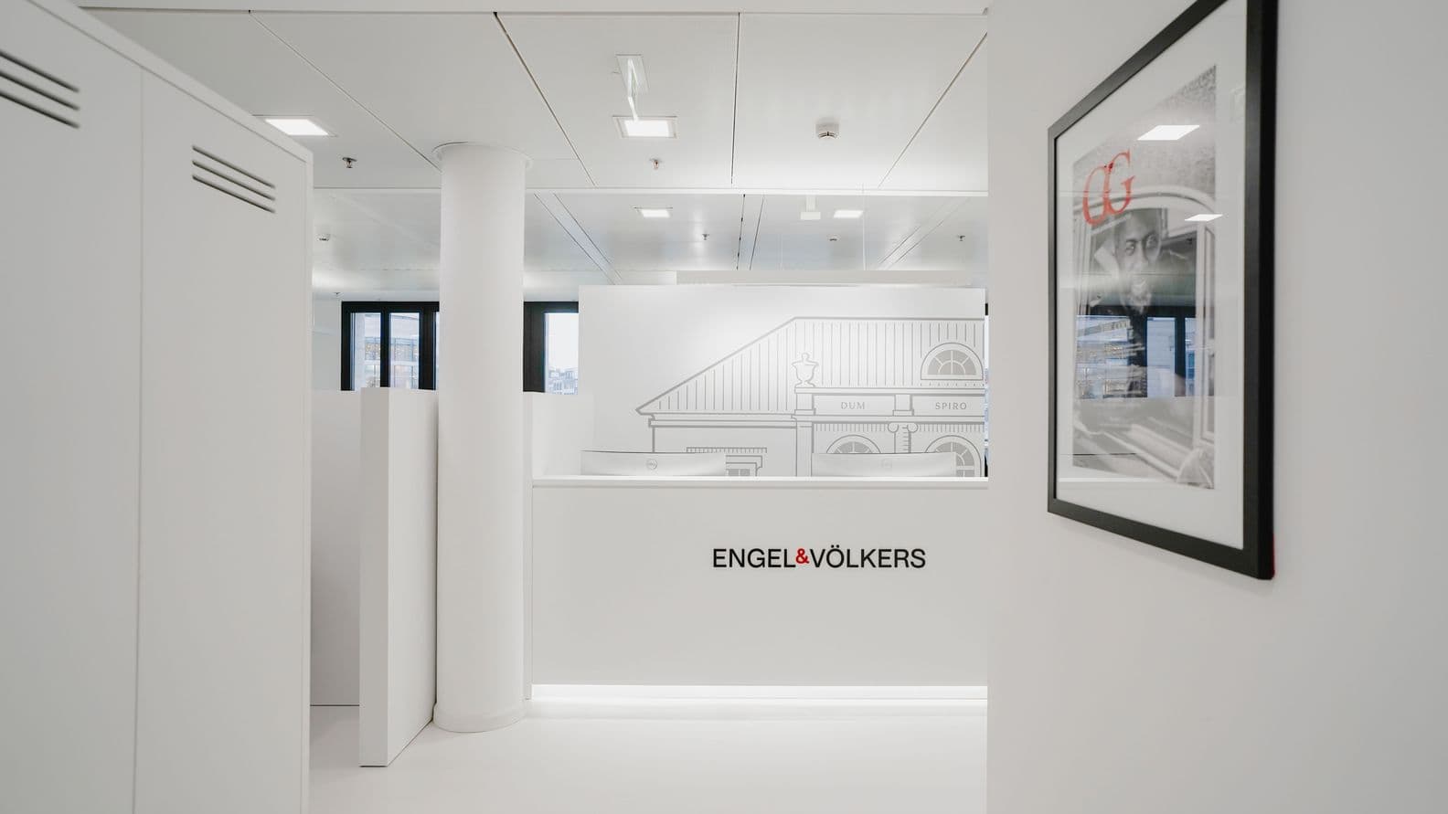 Moderne Büroeinrichtung mit weißen Wänden, einem gerahmten Bild und einem minimalistischen Design. Das „Engel & Völkers“-Logo prangt an prominenter Stelle.