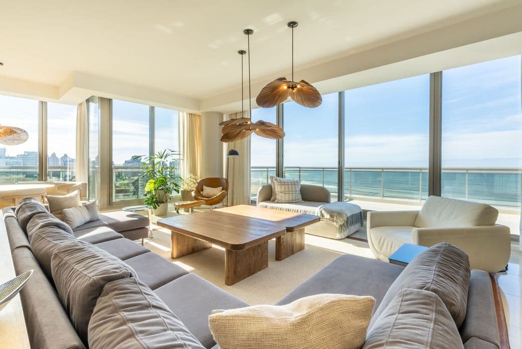 Sunlit modern living room with large windows overlooking the ocean, a gray sectional, wooden coffee table and sculptural pendant lights.