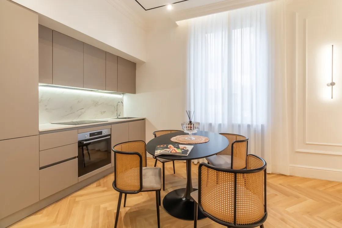 Modern kitchen with gray cabinetry, marble backsplash, built-in oven and a round black table with wicker chairs beside a large window.