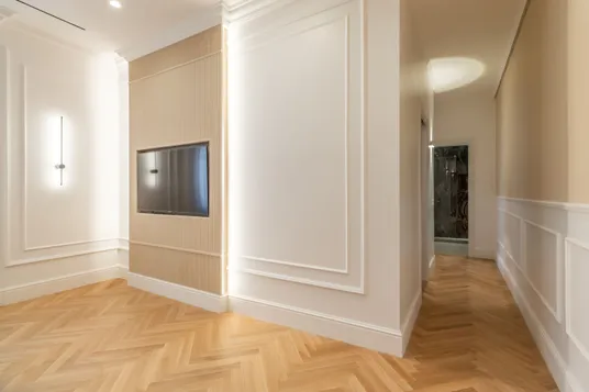 Corridoio luminoso con pareti bianche a pannelli e pannellatura beige scanalata con TV a schermo piatto; pavimento in legno caldo a spina di pesce e un'apertura che conduce a una stanza rivestita in marmo.