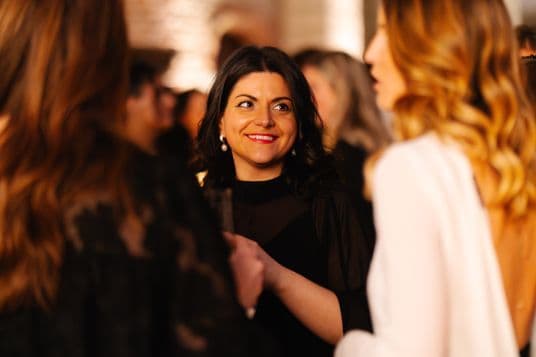 Centered woman with dark hair, wearing black, smiling with pearl earrings, among a warmly lit, blurred crowd at a social event.