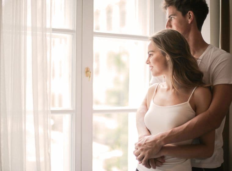 A couple embraces by a bright window with sheer curtains, looking out. The woman wears a light tank top, the man a white t-shirt.