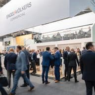 Eine belebte Messehalle mit dem Stand von Engel & Völkers Commercial auf zwei Ebenen, einer Glasbrüstung und einer Menschenmenge aus Fachleuten in Anzügen