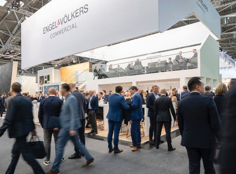 Eine belebte Messehalle mit dem Stand von Engel & Völkers Commercial auf zwei Ebenen, einer Glasbrüstung und einer Menschenmenge aus Fachleuten in Anzügen
