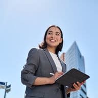 Low angle shot of a smiling woman in a gray suit holding a tablet and stylus, with city buildings and blue sky in the background.