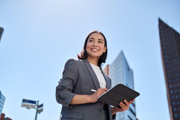 Low angle shot of a smiling woman in a gray suit holding a tablet and stylus, with city buildings and blue sky in the background.