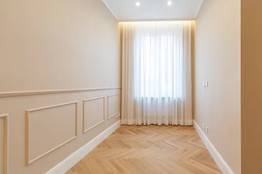Bright, minimalist hallway with beige walls, white half-height decorative wall panels, light herringbone wood flooring and a large window with sheer white curtains.