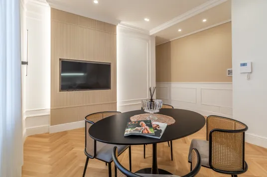 Modern living room with a round black dining table, Vienna straw-back chairs and a glass centerpiece; beige paneled wall with a wall-mounted TV, complemented by white wainscoting.